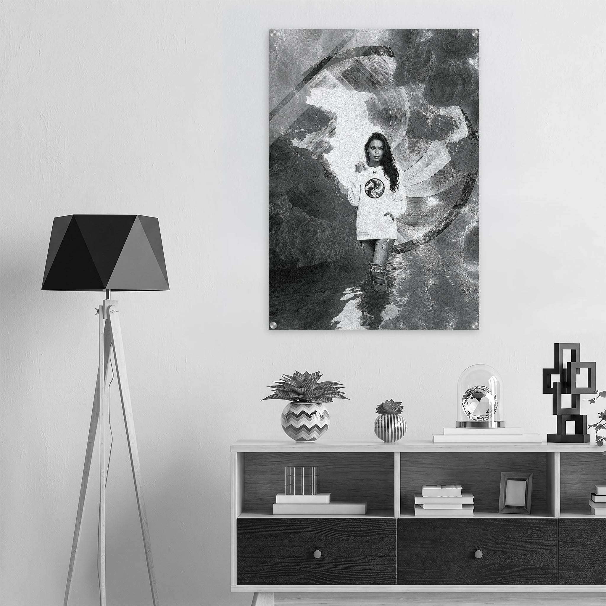 A black and white art print featuring a young girl in a white t-shirt standing in water, displayed on a light gray wall above a modern white and black sideboard with a black tripod floor lamp on the left.
