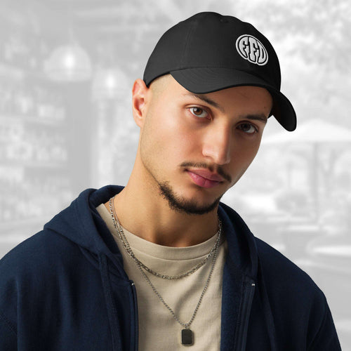 Man wearing the FFD13 x Under Armour Stamp Cap in black; the cap’s curved brim and white FFD monogram are prominent as he pairs it with a navy hoodie, beige T‑shirt and layered chain, against a softly blurred grey backdrop.