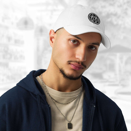 Man wearing the FFD14 x Under Armour Stamp Cap in white; the cap’s curved brim and black FFD monogram are prominent as he pairs it with a navy hoodie, beige T‑shirt and layered chain, against a softly blurred grey backdrop.