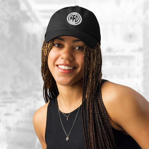 Front portrait of a smiling woman wearing the FFD15 x Under Armour Stamp Cap; the cap’s curved brim and bold white FFD monogram stand out above her long braids, with the background softly blurred.