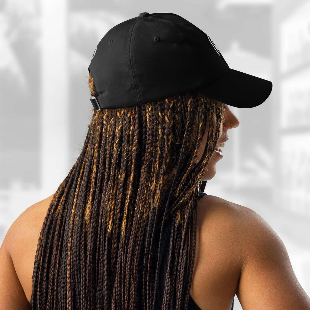 Back and side view of a woman with long braids wearing the FFD17 x Under Armour Stamp Cap; the adjustable fabric strap with a small white Under Armour logo patch and metal buckle is visible against a light blurred background.