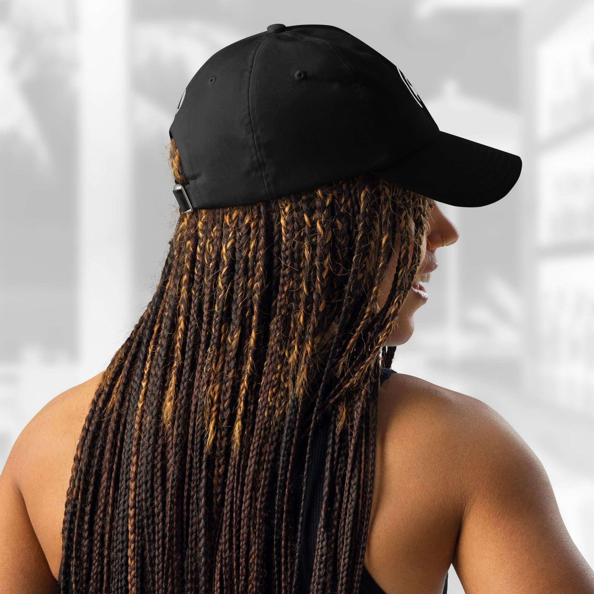 Back and side view of a woman with long braids wearing the FFD17 x Under Armour Stamp Cap; the adjustable fabric strap with a small white Under Armour logo patch and metal buckle is visible against a light blurred background.