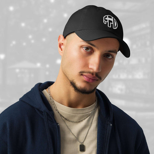 Man wearing the FFD5 x Under Armour Stamp Cap in black; the cap’s curved brim and white FFD monogram are prominent as he pairs it with a navy hoodie, beige T‑shirt and layered chain, against a softly blurred grey backdrop.