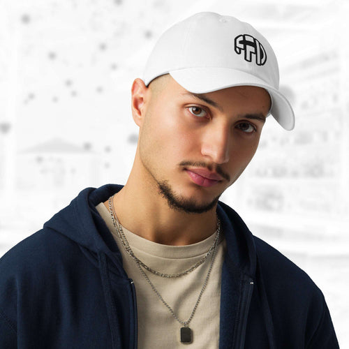 Man wearing the FFD6 x Under Armour Stamp Cap in white; the cap’s curved brim and black FFD monogram are prominent as he pairs it with a navy hoodie, beige T‑shirt and layered chain, against a softly blurred grey backdrop.