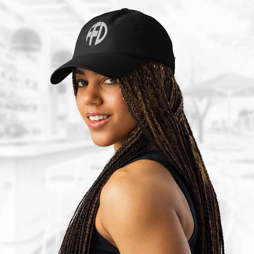 Three‑quarter portrait of a woman in the black FFD7 x Under Armour Stamp Cap; the white FFD emblem is clear on the front panel as her braids cascade over one shoulder against a soft, out‑of‑focus backdrop.