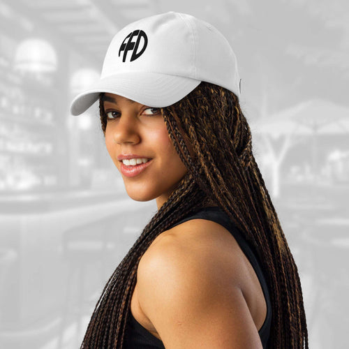 Three‑quarter portrait of a woman in the white FFD8 x Under Armour Stamp Cap; the black FFD emblem is clear on the front panel as her braids cascade over one shoulder against a soft, out‑of‑focus backdrop.