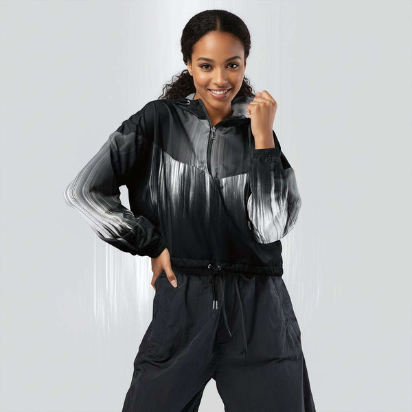 Female model wearing the HER Cropped Dither Distortion Windbreaker (No.02), three‑quarter front view. The cropped windbreaker has a black‑to‑white dither gradient pattern, long sleeves and a half‑zip front. The model holds the collar with one hand, the hood is down, and the jacket is paired with black trousers against a light grey background
