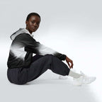 Side profile of a woman seated cross‑legged in the HER Cropped Dither Distortion Windbreaker (No.07). The cropped jacket shows a digital dither gradient from white to black, with the hood lowered and a drawcord at the hem. She wears black cargo trousers and white trainers, photographed against a neutral grey backdrop