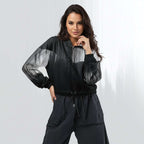 Female model wearing the HER Cropped Dither Distortion Windbreaker (No.10), three‑quarter front view. The cropped windbreaker has a black‑to‑white dither gradient pattern, long sleeves and a half‑zip front. The model holds the collar with one hand, the hood is down, and the jacket is paired with black trousers against a light grey background