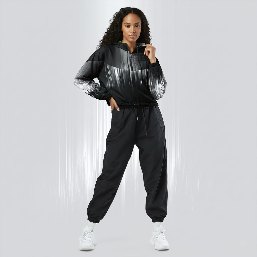 Full‑body view of a model standing in the HER Cropped Dither Distortion Windbreaker (No.02). The hooded jacket, with its abstract black‑to‑white gradient print and half‑zip closure, is worn with black trousers and white shoes. The scene has a minimalist grey background