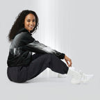 Side profile of a woman seated cross‑legged in the HER Cropped Dither Distortion Windbreaker (No.02). The cropped jacket shows a digital dither gradient from white to black, with the hood lowered and a drawcord at the hem. She wears black cargo trousers and white trainers, photographed against a neutral grey backdrop