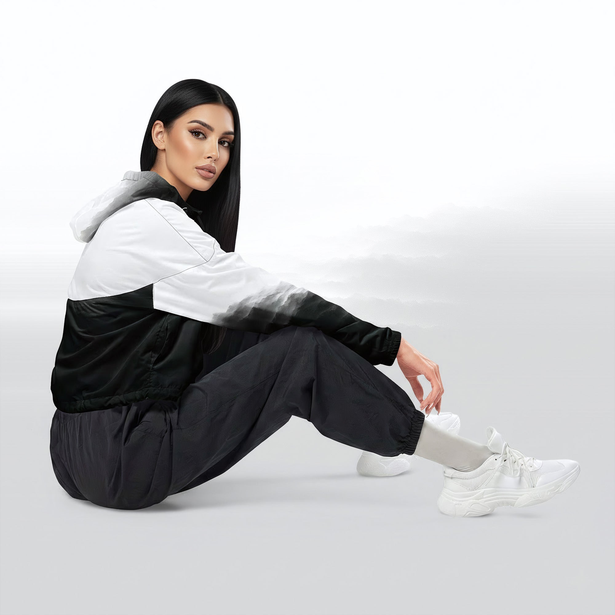 Side profile of a woman seated cross‑legged in the HER Cropped Dither Distortion Windbreaker (No.03). The cropped jacket shows a digital dither gradient from white to black, with the hood lowered and a drawcord at the hem. She wears black cargo trousers and white trainers, photographed against a neutral grey backdrop
