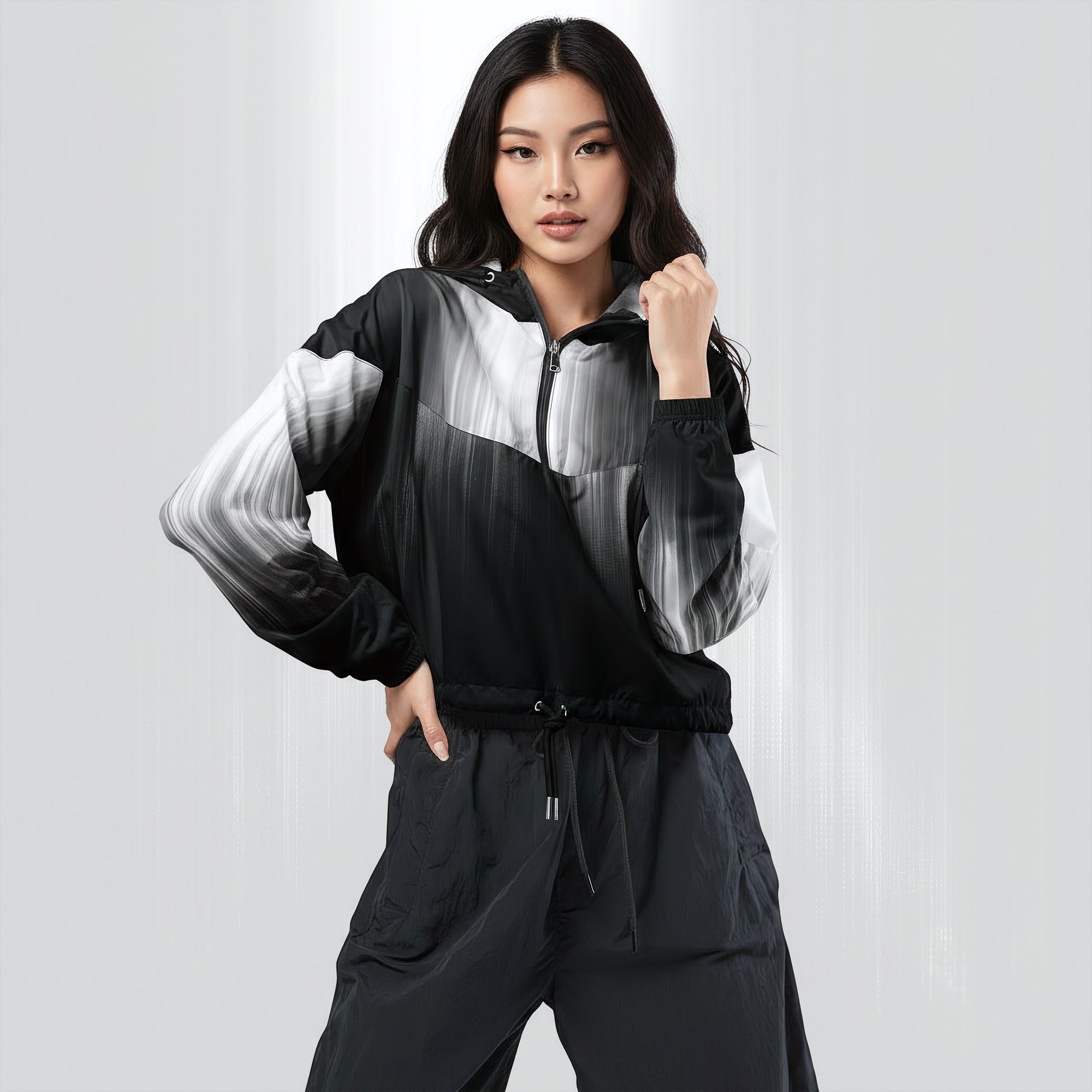 Female model wearing the HER Cropped Dither Distortion Windbreaker (No.04), three‑quarter front view. The cropped windbreaker has a black‑to‑white dither gradient pattern, long sleeves and a half‑zip front. The model holds the collar with one hand, the hood is down, and the jacket is paired with black trousers against a light grey background