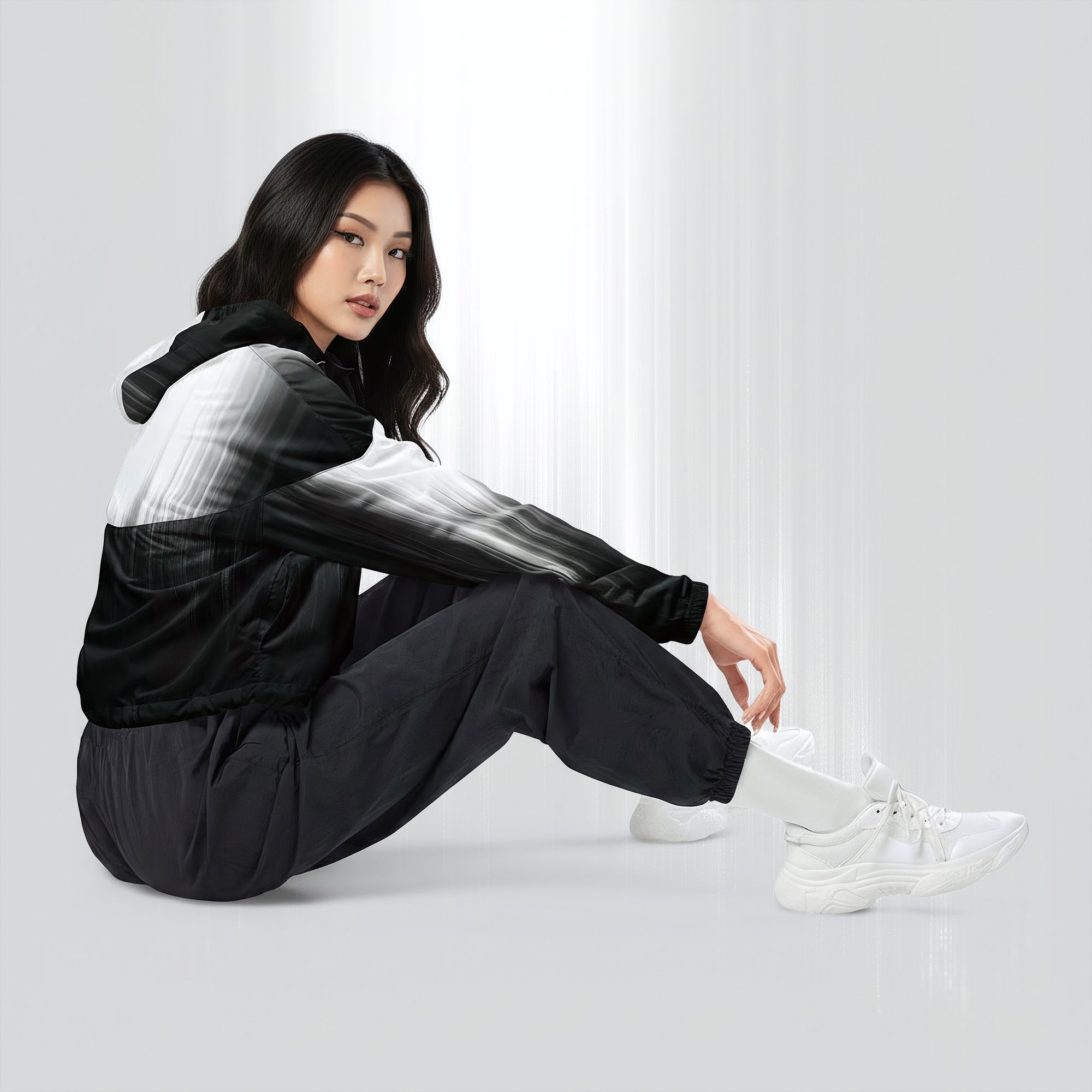 Side profile of a woman seated cross‑legged in the HER Cropped Dither Distortion Windbreaker (No.04). The cropped jacket shows a digital dither gradient from white to black, with the hood lowered and a drawcord at the hem. She wears black cargo trousers and white trainers, photographed against a neutral grey backdrop