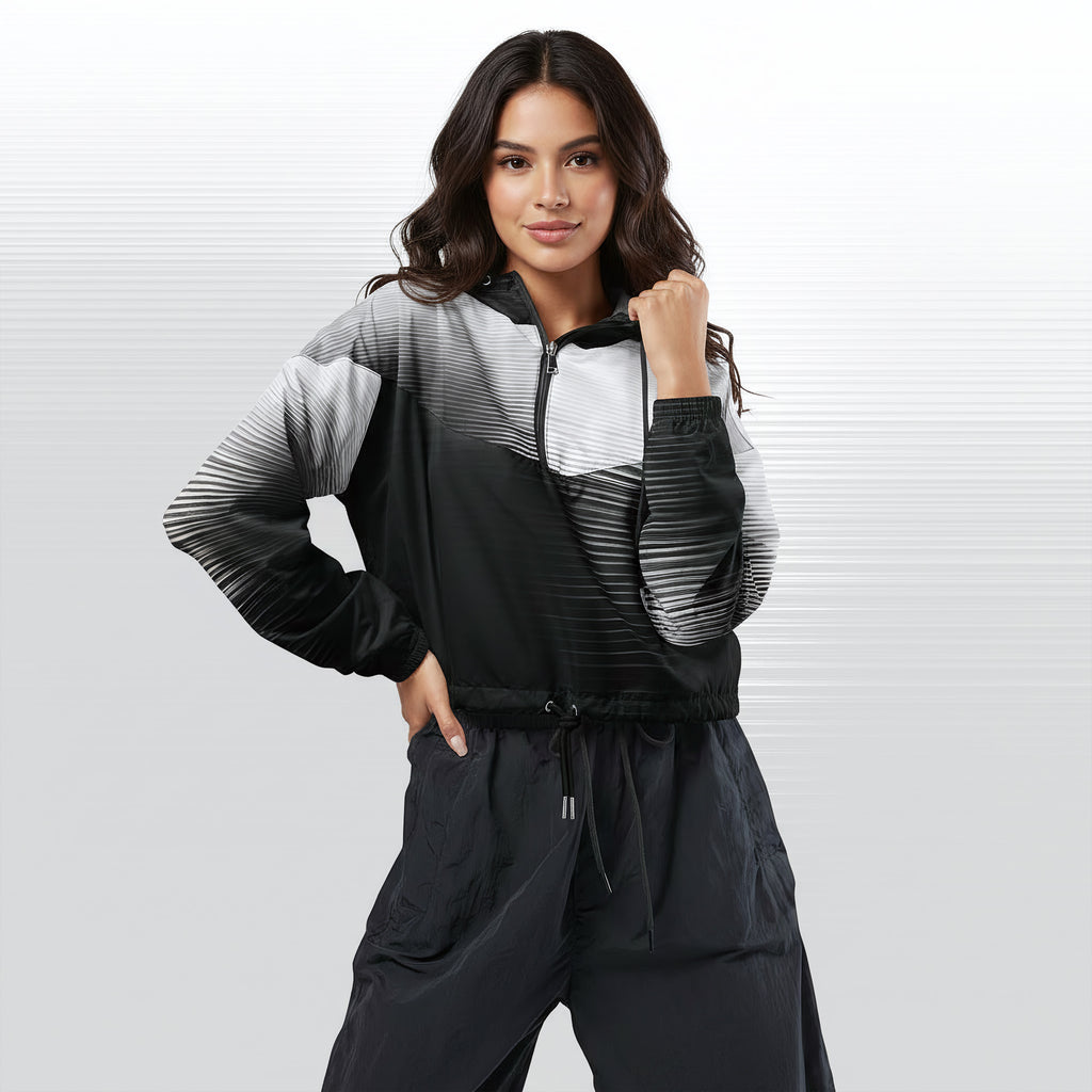 Female model wearing the HER Cropped Dither Distortion Windbreaker (No.05), three‑quarter front view. The cropped windbreaker has a black‑to‑white dither gradient pattern, long sleeves and a half‑zip front. The model holds the collar with one hand, the hood is down, and the jacket is paired with black trousers against a light grey background