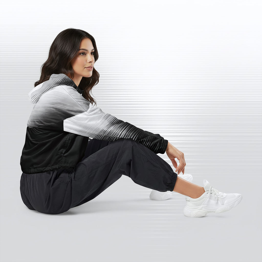 Side profile of a woman seated cross‑legged in the HER Cropped Dither Distortion Windbreaker (No.05). The cropped jacket shows a digital dither gradient from white to black, with the hood lowered and a drawcord at the hem. She wears black cargo trousers and white trainers, photographed against a neutral grey backdrop