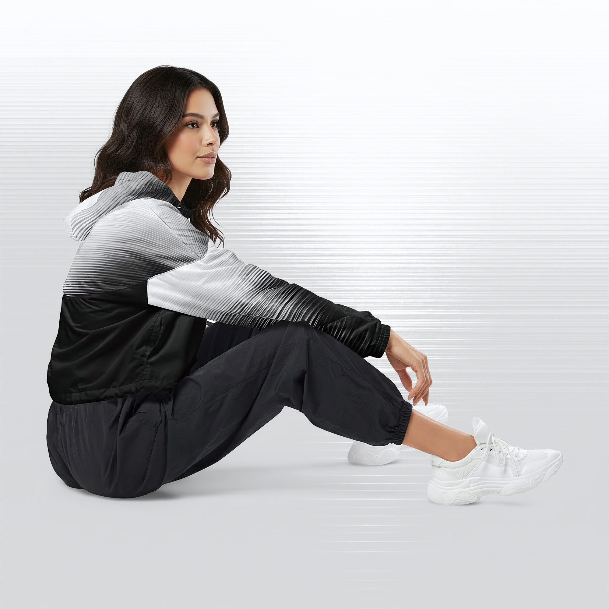 Side profile of a woman seated cross‑legged in the HER Cropped Dither Distortion Windbreaker (No.05). The cropped jacket shows a digital dither gradient from white to black, with the hood lowered and a drawcord at the hem. She wears black cargo trousers and white trainers, photographed against a neutral grey backdrop