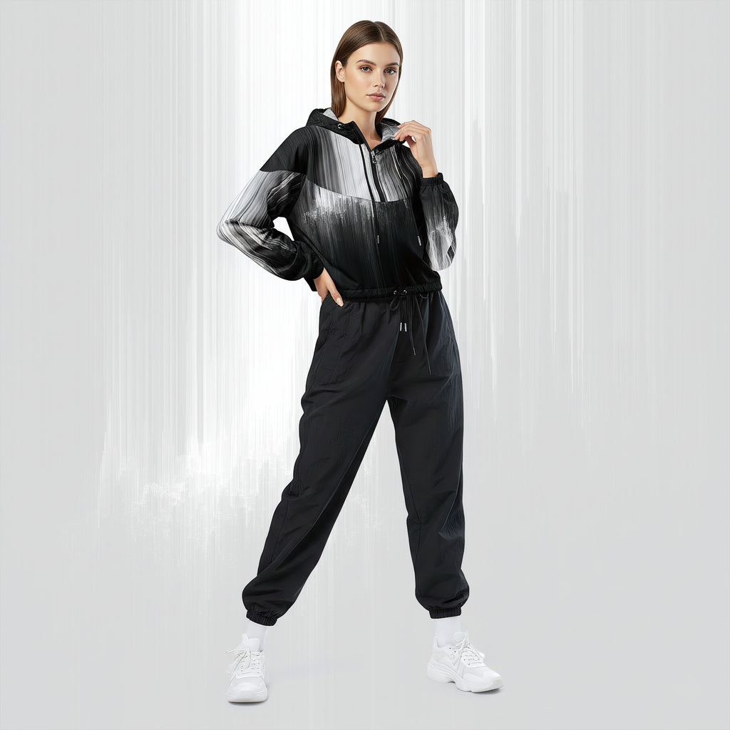 Full‑body view of a model standing in the HER Cropped Dither Distortion Windbreaker (No.06). The hooded jacket, with its abstract black‑to‑white gradient print and half‑zip closure, is worn with black trousers and white shoes. The scene has a minimalist grey background