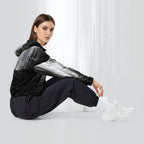Side profile of a woman seated cross‑legged in the HER Cropped Dither Distortion Windbreaker (No.06). The cropped jacket shows a digital dither gradient from white to black, with the hood lowered and a drawcord at the hem. She wears black cargo trousers and white trainers, photographed against a neutral grey backdrop