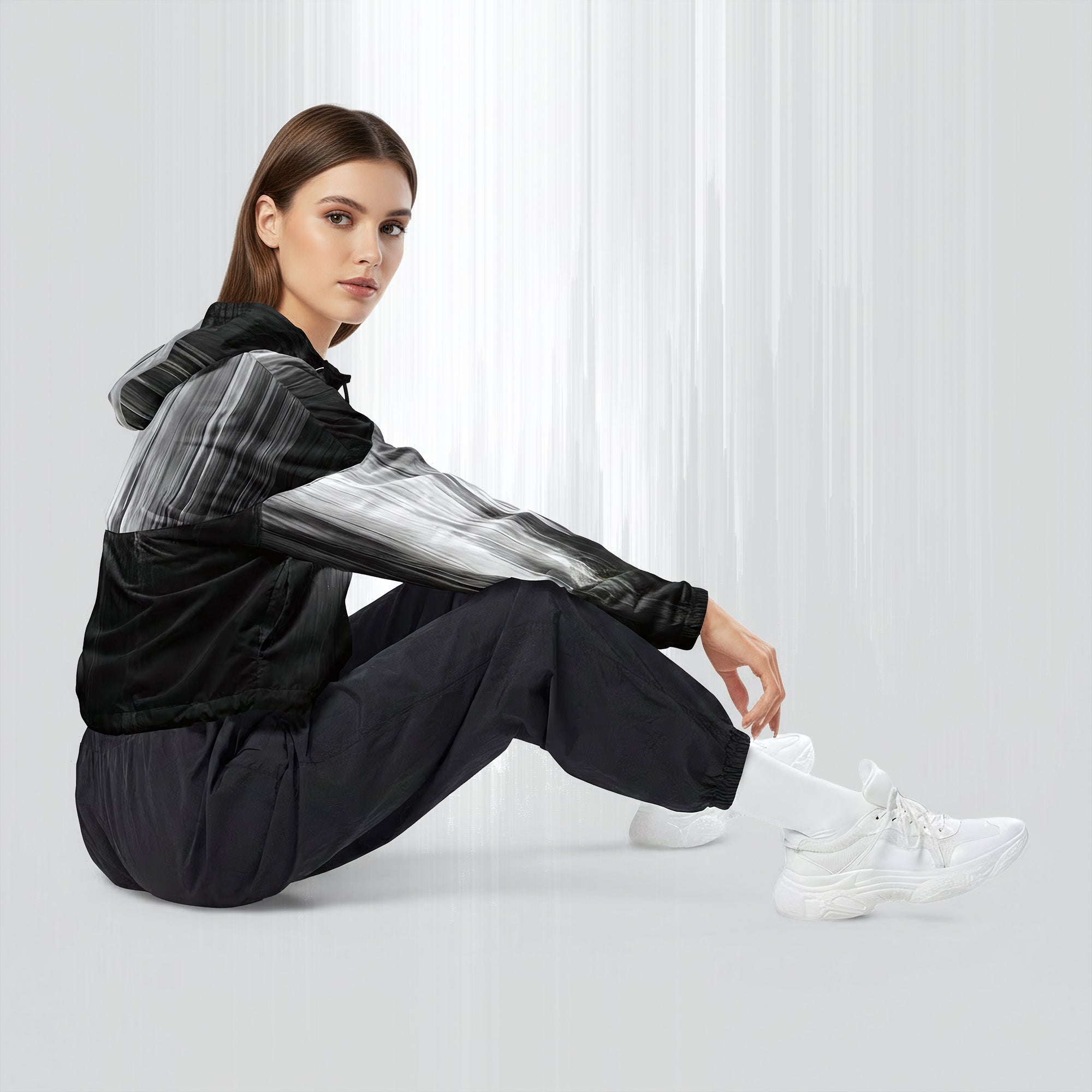 Side profile of a woman seated cross‑legged in the HER Cropped Dither Distortion Windbreaker (No.06). The cropped jacket shows a digital dither gradient from white to black, with the hood lowered and a drawcord at the hem. She wears black cargo trousers and white trainers, photographed against a neutral grey backdrop