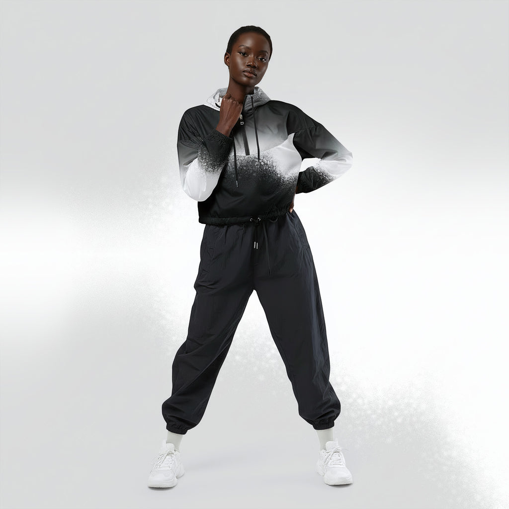 Full‑body view of a model standing in the HER Cropped Dither Distortion Windbreaker (No.07). The hooded jacket, with its abstract black‑to‑white gradient print and half‑zip closure, is worn with black trousers and white shoes. The scene has a minimalist grey background