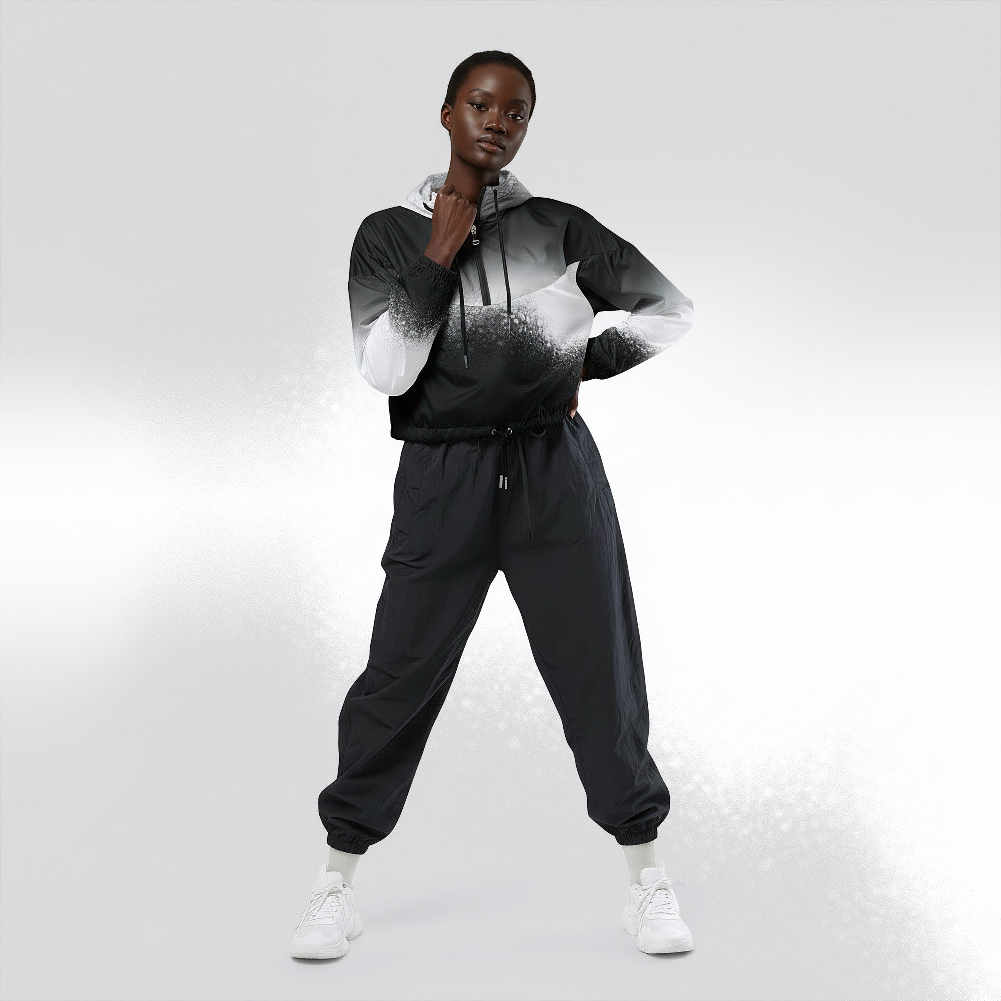Full‑body view of a model standing in the HER Cropped Dither Distortion Windbreaker (No.07). The hooded jacket, with its abstract black‑to‑white gradient print and half‑zip closure, is worn with black trousers and white shoes. The scene has a minimalist grey background