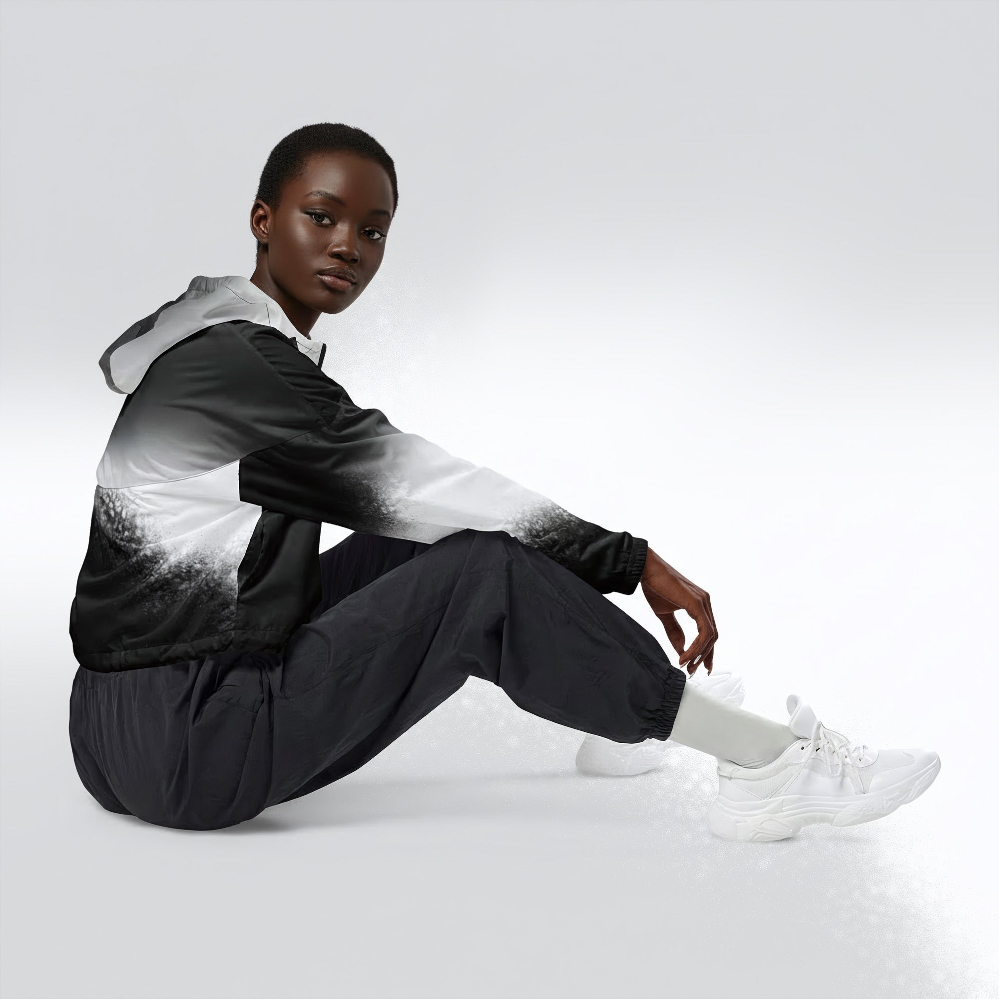 Side profile of a woman seated cross‑legged in the HER Cropped Dither Distortion Windbreaker (No.07). The cropped jacket shows a digital dither gradient from white to black, with the hood lowered and a drawcord at the hem. She wears black cargo trousers and white trainers, photographed against a neutral grey backdrop