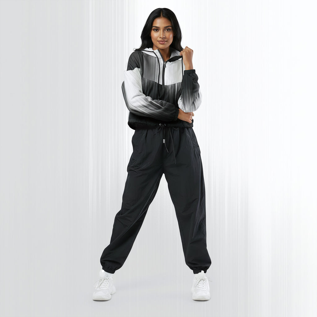 Full‑body view of a model standing in the HER Cropped Dither Distortion Windbreaker (No.08). The hooded jacket, with its abstract black‑to‑white gradient print and half‑zip closure, is worn with black trousers and white shoes. The scene has a minimalist grey background
