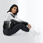 Side profile of a woman seated cross‑legged in the HER Cropped Dither Distortion Windbreaker (No.08). The cropped jacket shows a digital dither gradient from white to black, with the hood lowered and a drawcord at the hem. She wears black cargo trousers and white trainers, photographed against a neutral grey backdrop