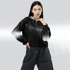 Female model wearing the HER Cropped Dither Distortion Windbreaker (No.09), three‑quarter front view. The cropped windbreaker has a black‑to‑white dither gradient pattern, long sleeves and a half‑zip front. The model holds the collar with one hand, the hood is down, and the jacket is paired with black trousers against a light grey background