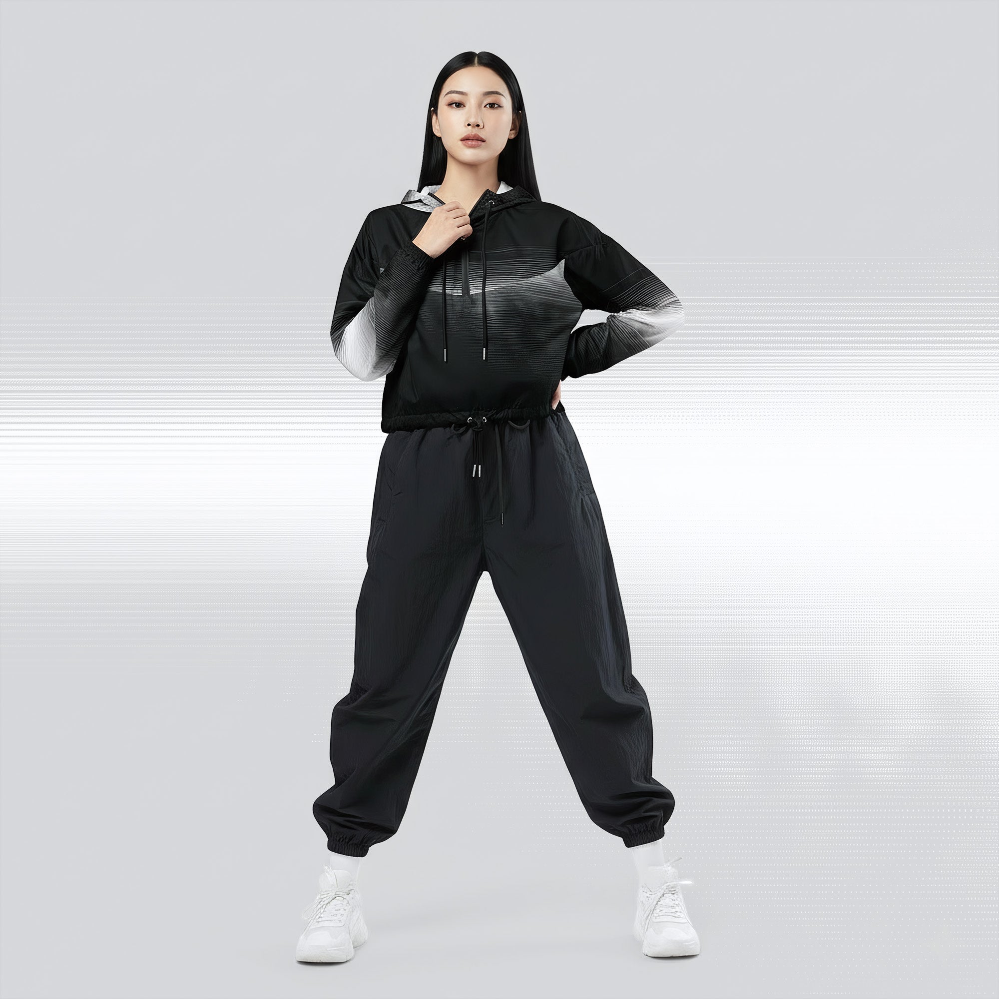 Full‑body view of a model standing in the HER Cropped Dither Distortion Windbreaker (No.09). The hooded jacket, with its abstract black‑to‑white gradient print and half‑zip closure, is worn with black trousers and white shoes. The scene has a minimalist grey background