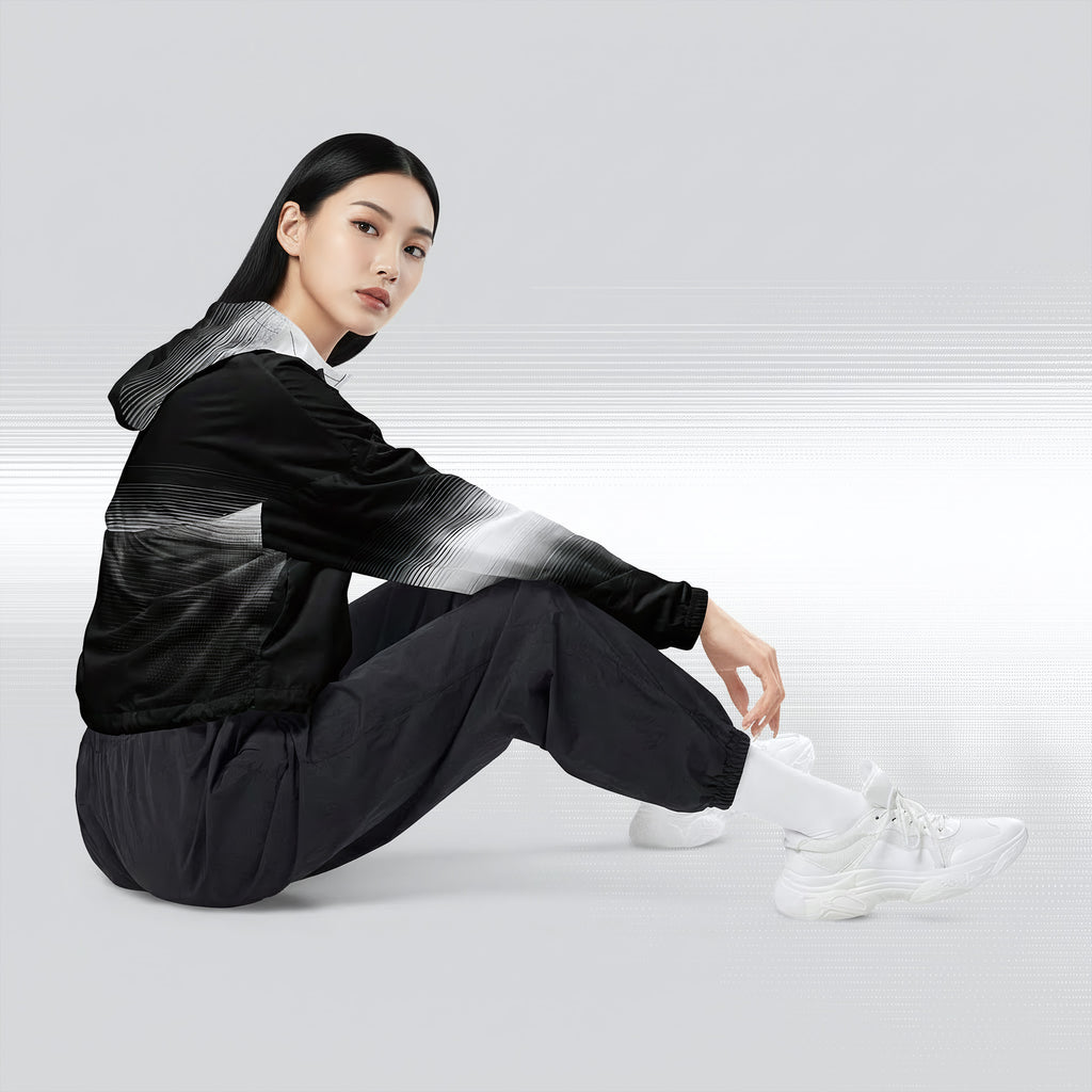 Side profile of a woman seated cross‑legged in the HER Cropped Dither Distortion Windbreaker (No.09). The cropped jacket shows a digital dither gradient from white to black, with the hood lowered and a drawcord at the hem. She wears black cargo trousers and white trainers, photographed against a neutral grey backdrop