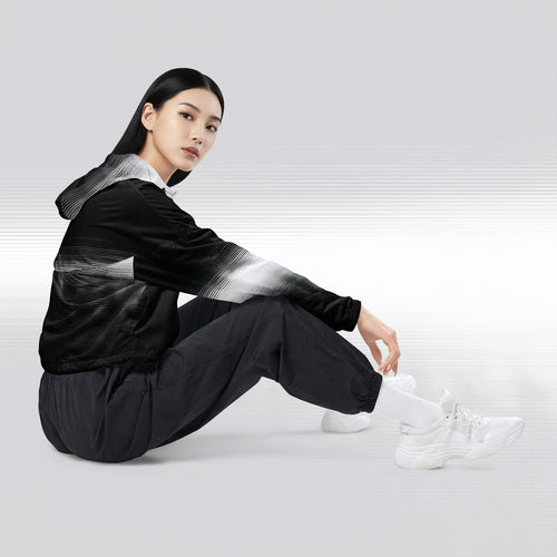 Side profile of a woman seated cross‑legged in the HER Cropped Dither Distortion Windbreaker (No.09). The cropped jacket shows a digital dither gradient from white to black, with the hood lowered and a drawcord at the hem. She wears black cargo trousers and white trainers, photographed against a neutral grey backdrop