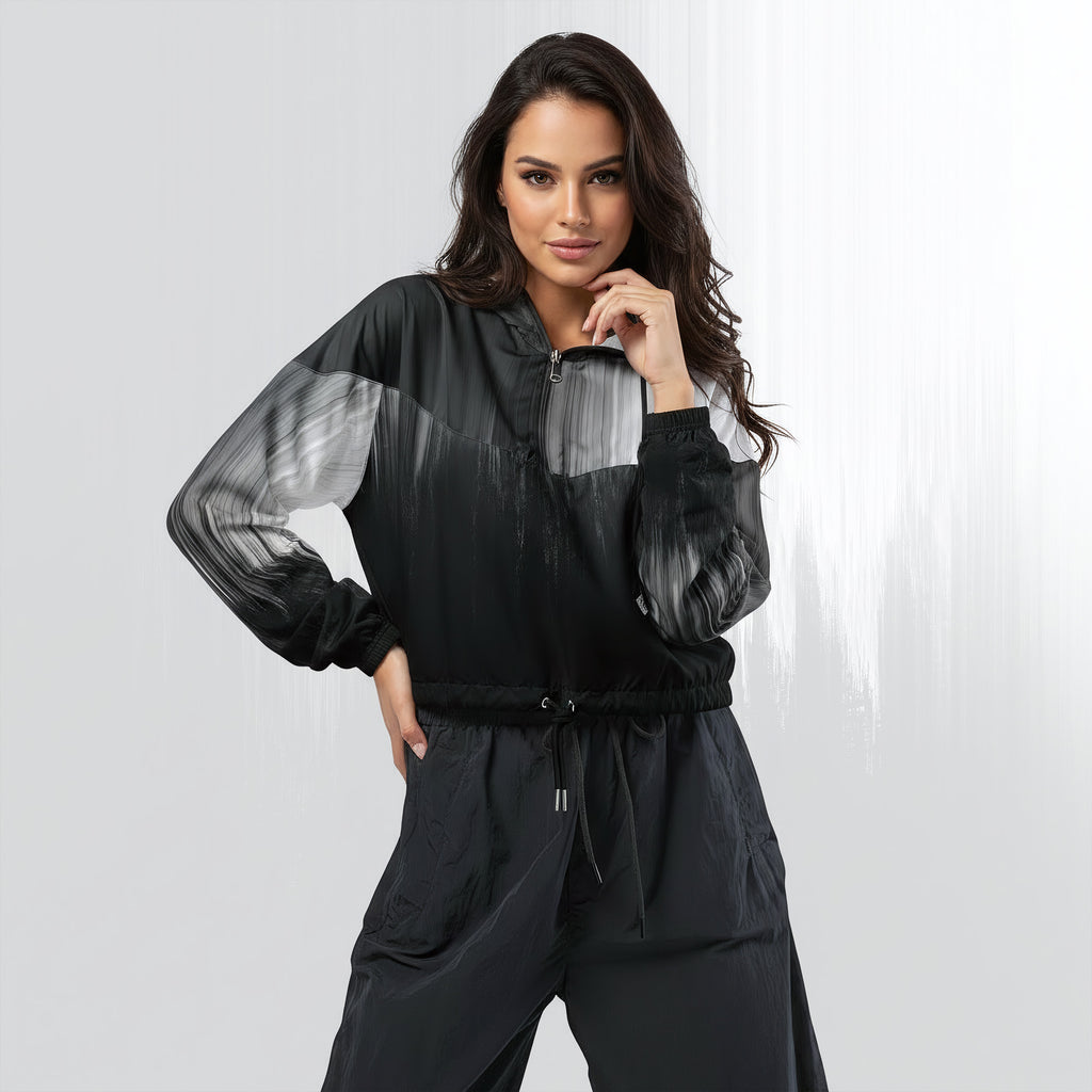 Female model wearing the HER Cropped Dither Distortion Windbreaker (No.10), three‑quarter front view. The cropped windbreaker has a black‑to‑white dither gradient pattern, long sleeves and a half‑zip front. The model holds the collar with one hand, the hood is down, and the jacket is paired with black trousers against a light grey background