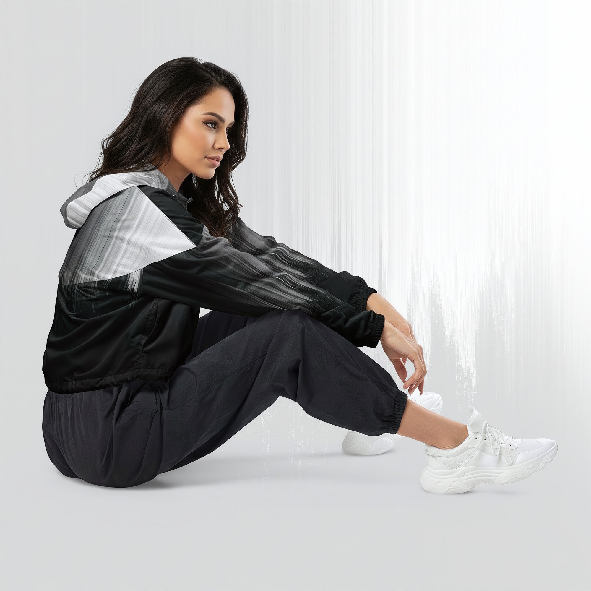 Side profile of a woman seated cross‑legged in the HER Cropped Dither Distortion Windbreaker (No.10). The cropped jacket shows a digital dither gradient from white to black, with the hood lowered and a drawcord at the hem. She wears black cargo trousers and white trainers, photographed against a neutral grey backdrop