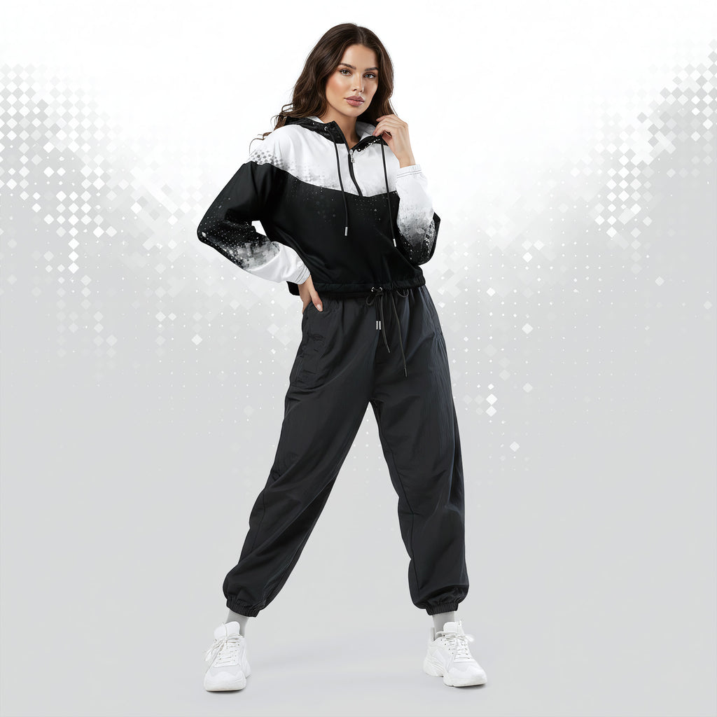 A woman modeling a black and white color-blocked windbreaker jacket with matching black jogger pants and white sneakers, posed against a light gray studio background with a subtle dotted pattern.