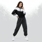 A woman modeling a black and white color-blocked windbreaker jacket with matching black jogger pants and white sneakers, posed against a light gray studio background with a subtle dotted pattern.
