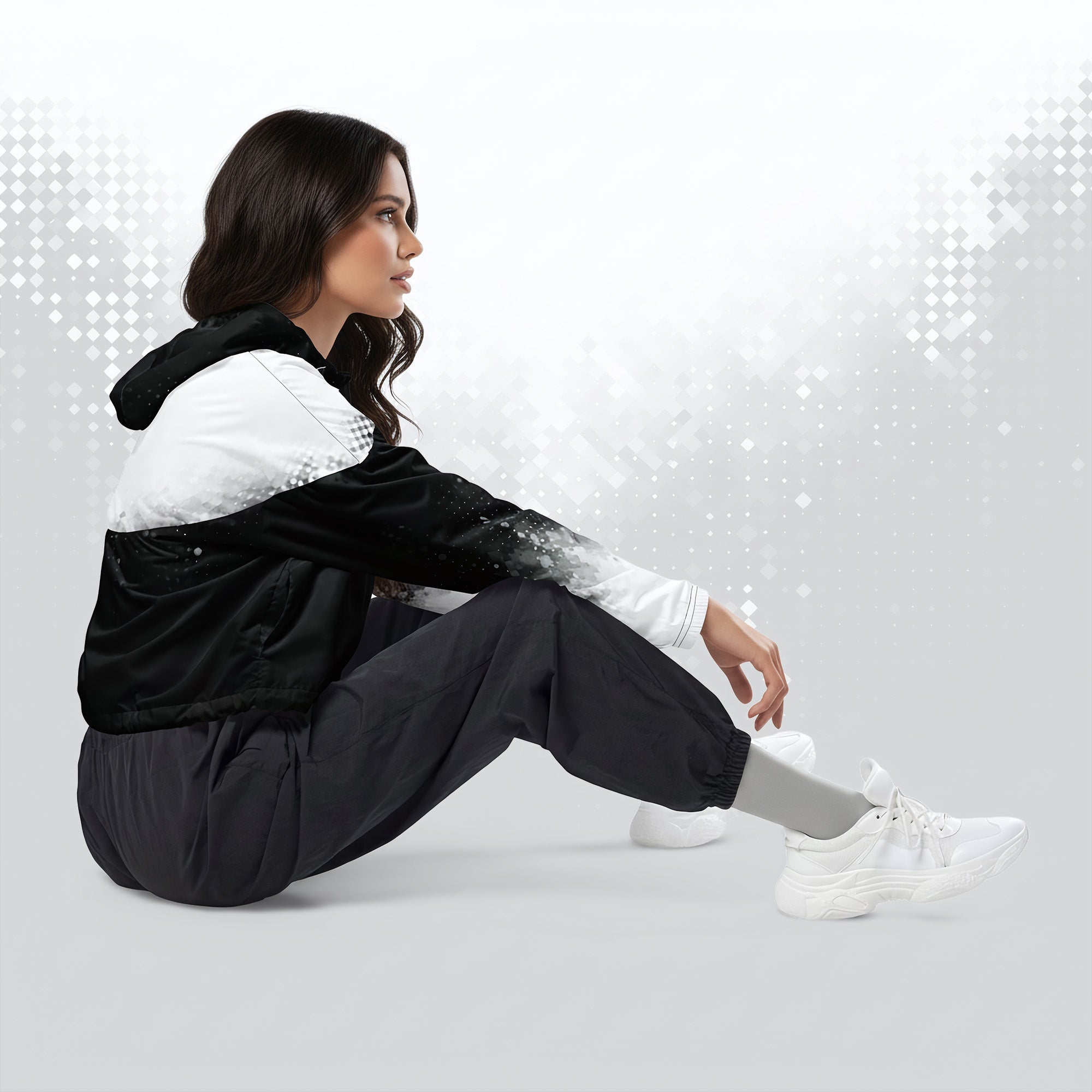 A woman sits on the floor in a studio setting, wearing a black and white hooded jacket, loose black pants, and white sneakers against a light gray background with subtle digital bokeh effects.
