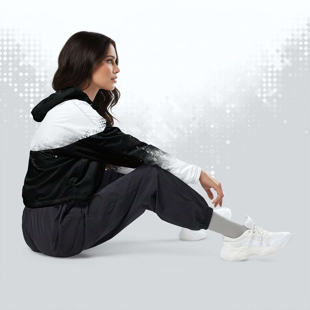 A woman sits on the floor in a studio setting, wearing a black and white hooded jacket, loose black pants, and white sneakers against a light gray background with subtle digital bokeh effects.