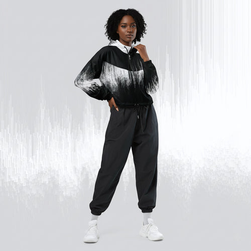 A model wearing a black and white abstract-patterned zip-up hoodie with black jogger pants and white sneakers, standing against a light gray background with vertical white streaks.
