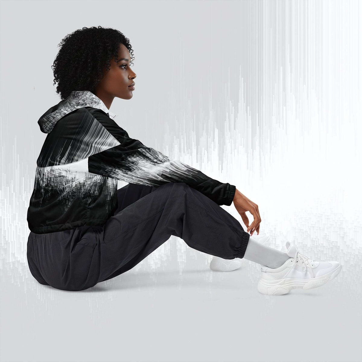 A model sits on the floor in profile, wearing a black and white abstract brushstroke print hooded jacket, black jogger pants, and white sneakers against a light gray studio background with vertical light streaks.