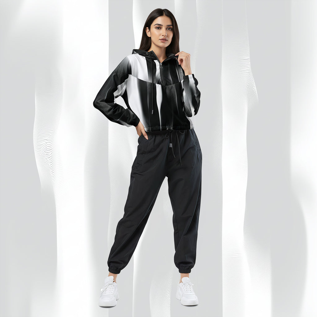 A woman modeling a black, white, and gray color-blocked zip-up hoodie with matching black jogger pants and white sneakers, set against a white background with soft vertical shadows.