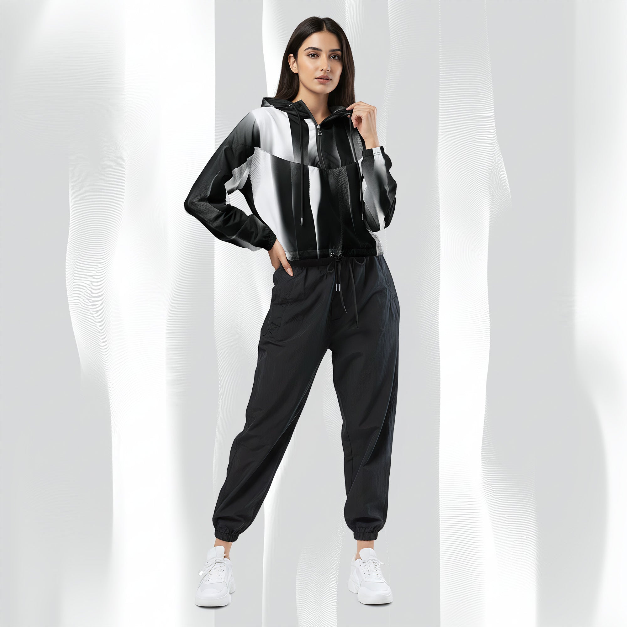 A woman modeling a black, white, and gray color-blocked zip-up hoodie with matching black jogger pants and white sneakers, set against a white background with soft vertical shadows.
