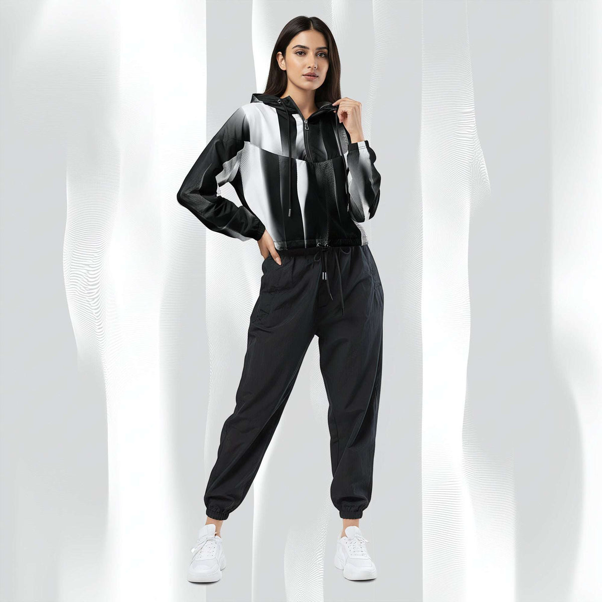 A woman modeling a black, white, and gray color-blocked zip-up hoodie with matching black jogger pants and white sneakers, set against a white background with soft vertical shadows.