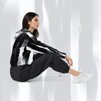 A woman sits on a white floor wearing a black and white striped hooded jacket, black jogger pants, and white sneakers in a bright, modern studio setting with soft draped light.