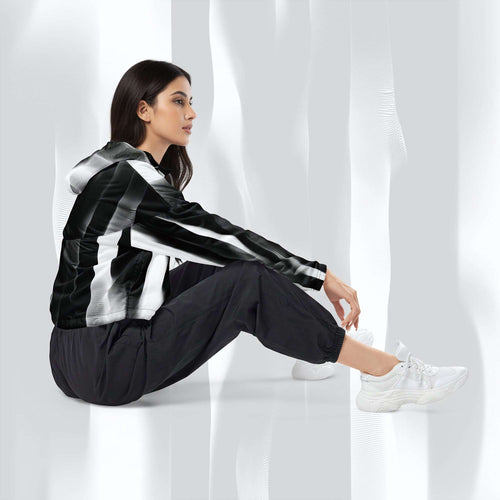A woman sits on a white floor wearing a black and white striped hooded jacket, black jogger pants, and white sneakers in a bright, modern studio setting with soft draped light.