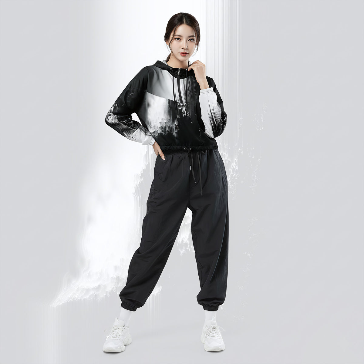 A woman modeling a black and white graphic hoodie with matching black jogger sweatpants and white sneakers, standing against a light gray studio background with a stylized white smoke effect.