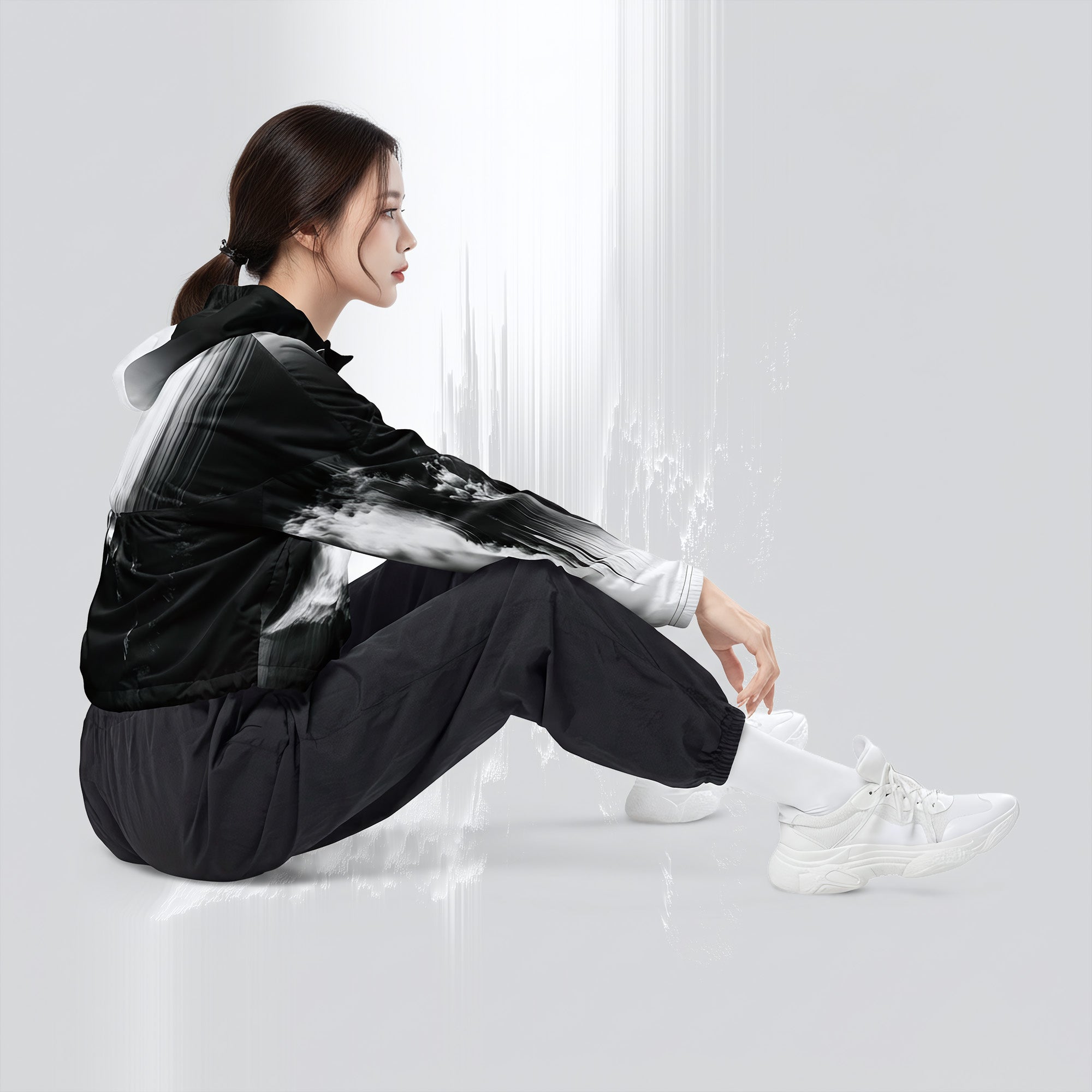 A woman sits on the floor wearing a black and white hooded jacket with a brushstroke pattern, black pants, and white sneakers in a bright, minimalist studio setting.