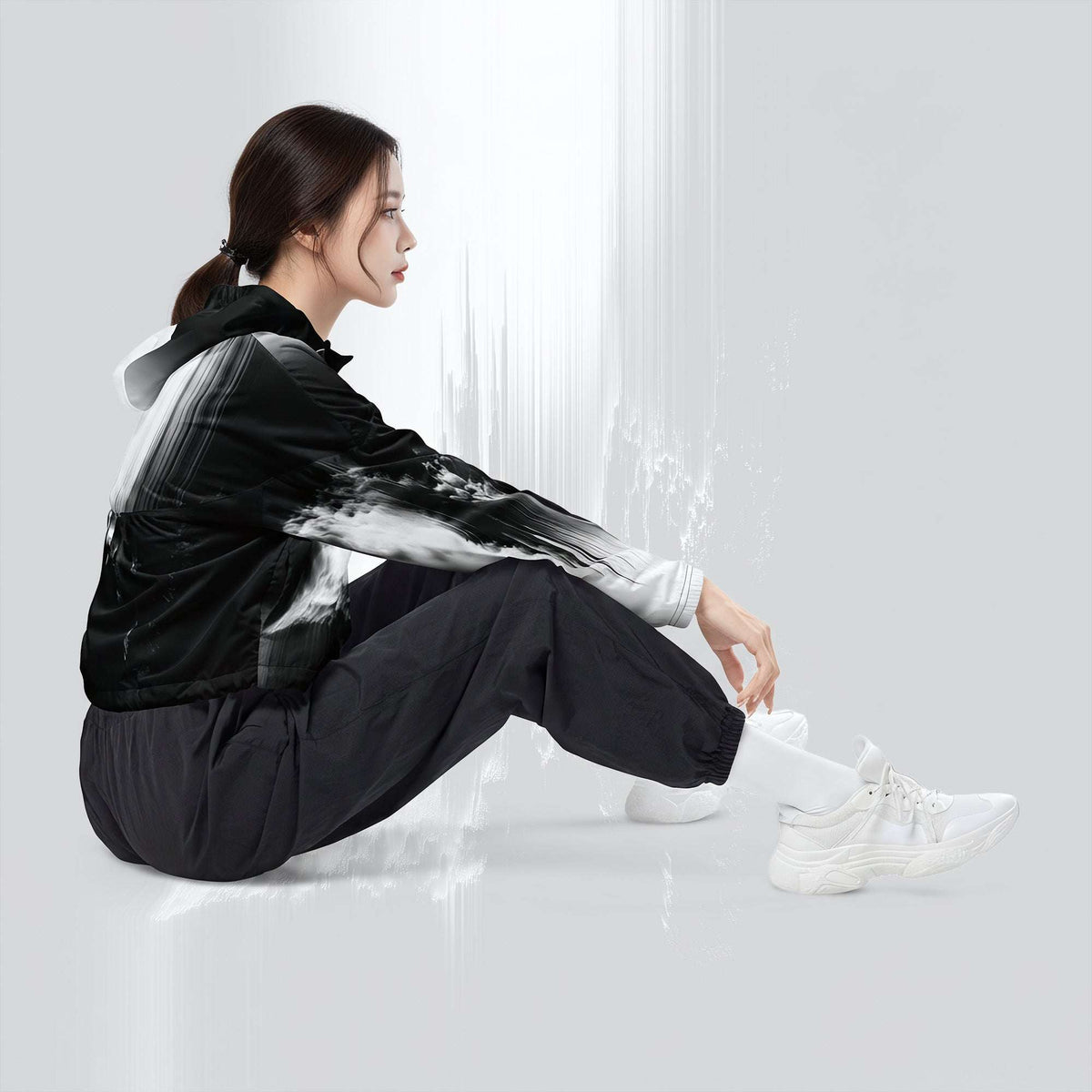 A woman sits on the floor wearing a black and white hooded jacket with a brushstroke pattern, black pants, and white sneakers in a bright, minimalist studio setting.