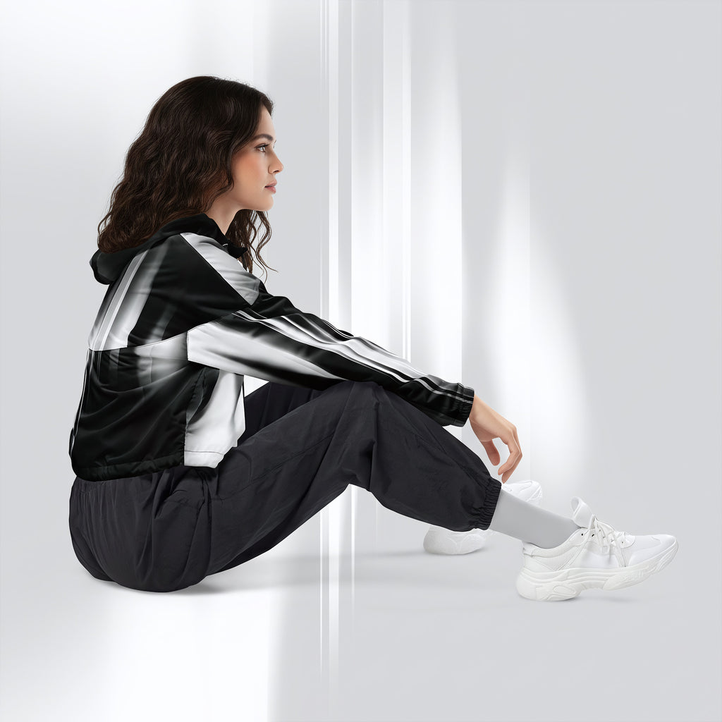 A woman sits on a white floor against a white wall, wearing a black and white striped hooded jacket, black jogger pants, white socks, and white sneakers.